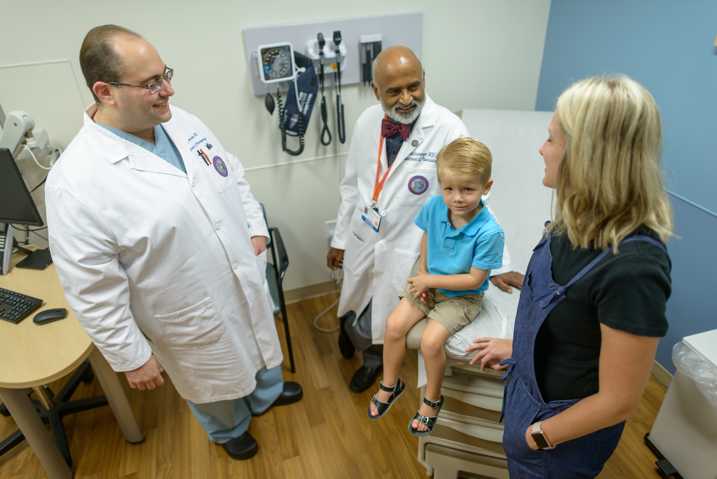 Two UF Health pediatric neurosurgeons speak with the mother of a young male patient as he sits on an examination chair.