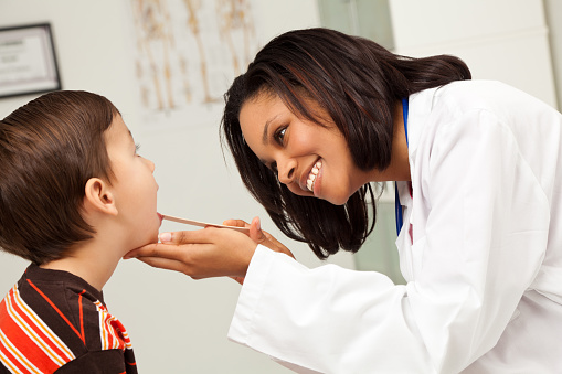 A female otolaryngologist uses a tongue depressor to push down on a young boys tongue and look into his mouth.