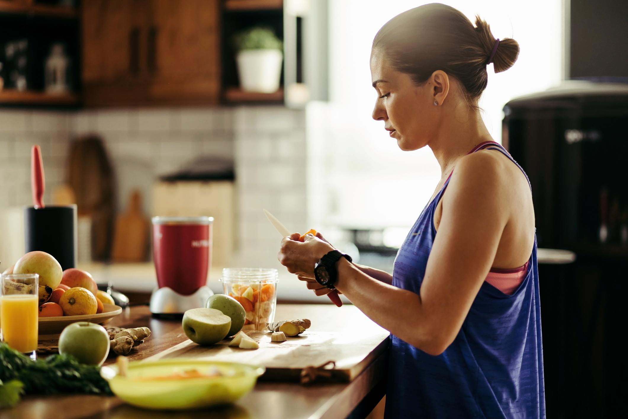 woman in athletic clothing standing at a kitchen counter cutting up fresh fruit for a smoothie