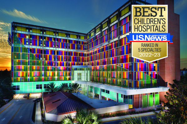 UF Health Shands Children's Hospital is backlit by the setting sun. Lights under the palms and trees in front of the building light up the sea green reflective glass and rectangular blue, yellow, red, and purple screens that cover the windows of the building. 