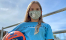 Kendall Lewis stands next to a volleyball net on a sunny day. She is wearing a face mask and is carrying an orange and blue Florida Gators volleyball. 